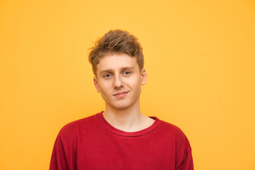 Wall Mural - Handsome young man in a red sweatshirt stands on a yellow background, looks at the camera and smiles. Portrait of a happy teenager on a yellow background.