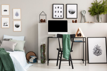 Green cloth on black wooden chair at desk with computer, wooden triangle and cones, real photo with gallery of posters on empty grey wall