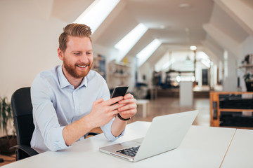 Wall Mural - Positive ginger businessman using smartphone while sitting at the desk with laptop.