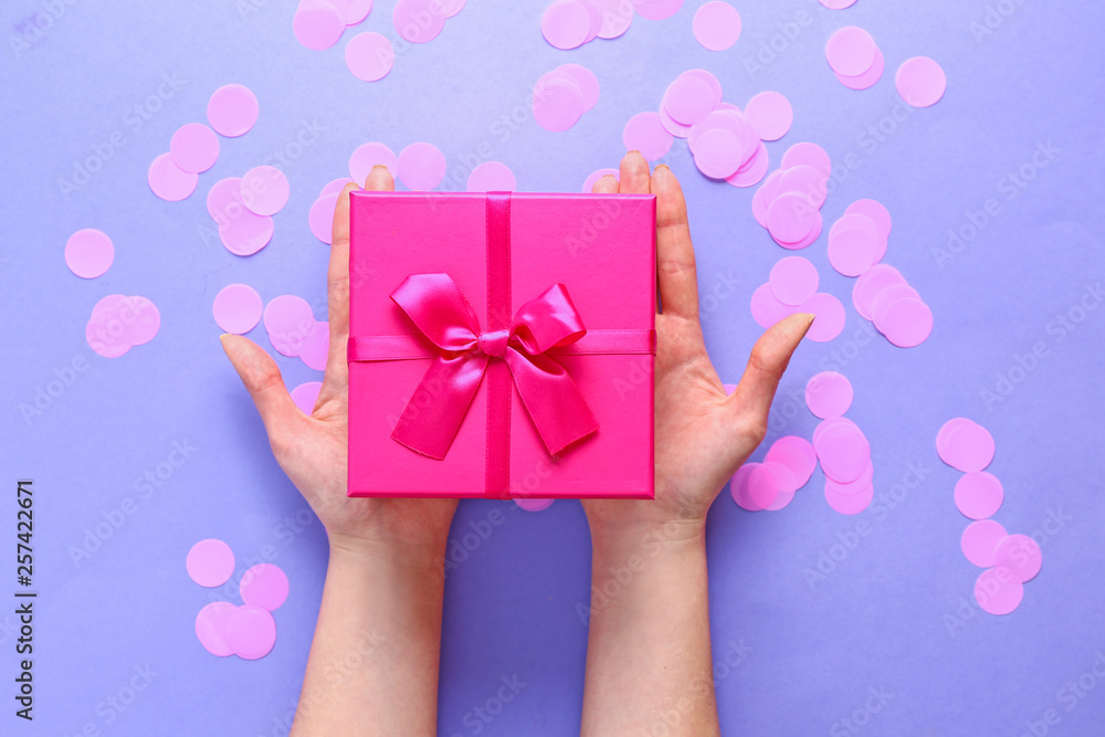Female hands with gift box and confetti on color background