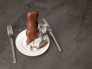 Partially unwrapped yummy chocolate Easter bunny standing on a plate, waiting to be eaten with knife and fork; studio stillife shot with copy space 