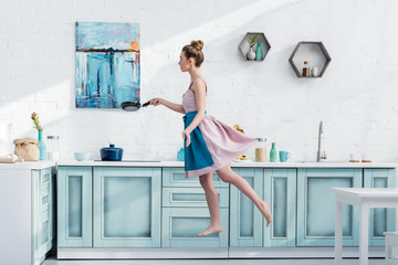 attractive barefoot young woman in apron levitating in kitchen while holding pan