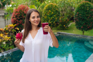 Wall Mural - Portrait of happy girl with beautiful smile holds glass of dragon fruit smoothie and fruit in her hands background of pool