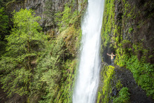 Columbia River Gorge, OR, USA. A Man Stands At The Opening Of A Tunnel Along A Lush Green, Moss-lined Trail With A Waterfall Falling From Above.