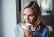© Westend61 - Smiling woman holding a cup of coffee at the window at home