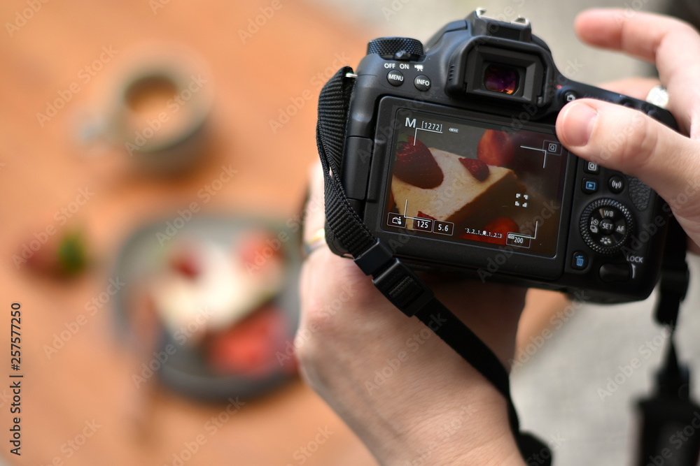 Hands of a photographer holding a camera, touching the screen with one finger taking photo of a cake on the plate and a cup of coffee in the blurred background. Closeup. - obrazy, fototapety, plakaty 