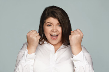 Wall Mural - Picture of beautiful overjoyed ecstatic young dark haired plus size female employee in white shirt screaming excitedly and clenching fists, happy ro receive promotion for hard work. Selective focus