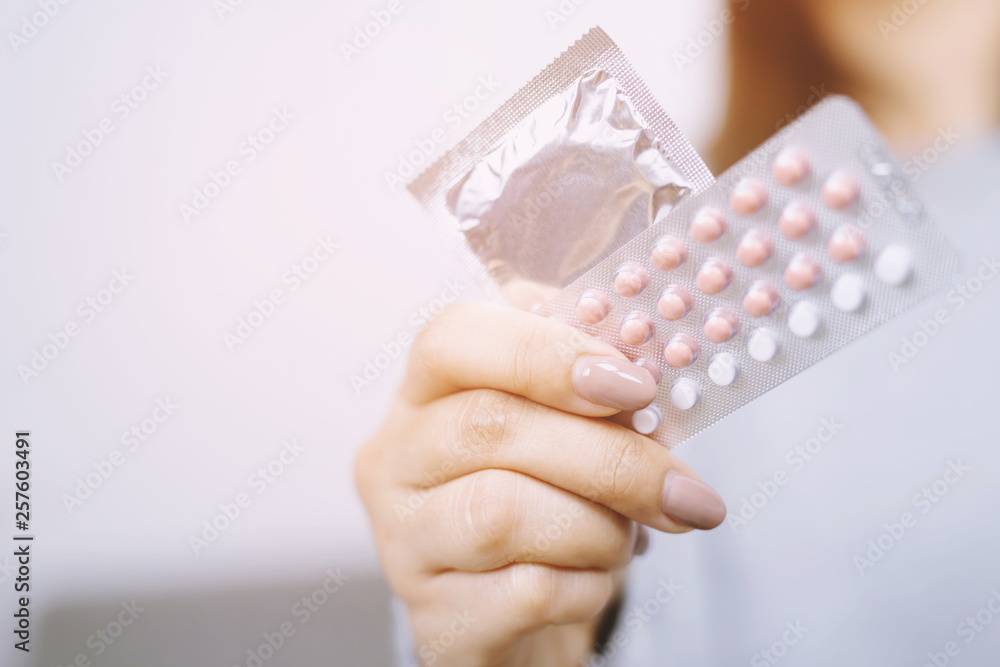 Contraceptive means: Woman hand holding contraceptive pills and condom sitting on sofa background. Protection, safe sex. Contraception, concept birth control. Copy space - obrazy, fototapety, plakaty 