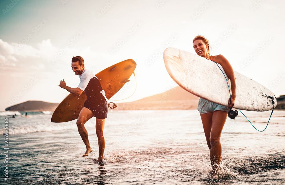 Happy surfer couple running with surfboards along the sea shore - Sporty people having fun going to surf together at sunset - Extreme surfing sport and youth relationship lifestyle concept - obrazy, fototapety, plakaty 