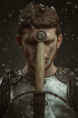 Emotional portrait of a young man in knight armor and a sword against a dark background.