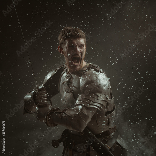 Fotomural Emotional portrait of a young man in knight armor and a sword against a dark background