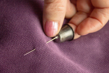 White elderly woman holding a needle on the middle finger wearing a thimble, the needle pierces the lilac textured fabric