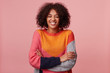 Happy joyful attractive African American with an afro hairstyle closed eyes and laughs from something funny, stand with arms crossed, wearing colorful sweater, isolated on pink