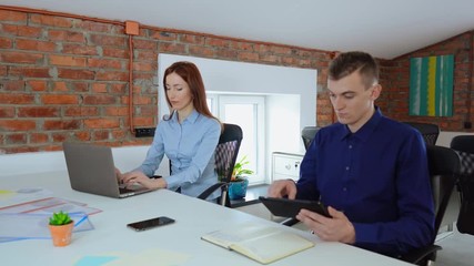 Poster - two young employees in startup company sitting at the desk in office with casual interior. redhead businesswoman typing on laptop businessman holding digital tablet