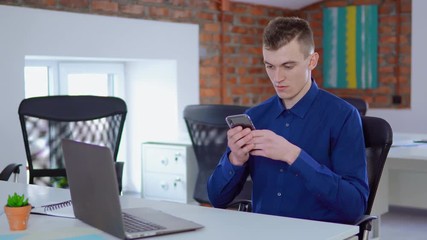 Poster - young worker in startup company using smartphone texting message or checking email sitting at the white desk in loft office businessman at work daytime