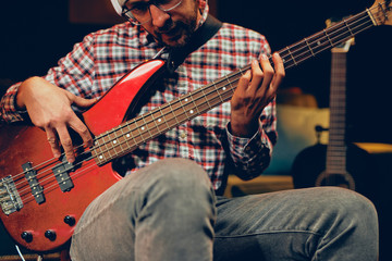 Wall Mural - Young Caucasian man with hat on head playing bass guitar while sitting in home studio.