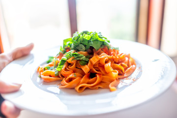 Closeup of hands waiter serving fresh red tomato sauce tagliatelle pasta corn gluten free noodles on plate in marinara with chopped basil greens in restaurant on white plate