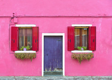 Beautiful Facade With Red Shutters And  Flowers And Purple Door On Pink Wall. Colorful Houses On Burano Island Near Venice, Italy