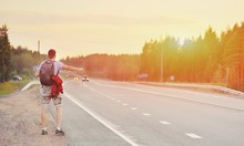 Hitchhiking Young Traveller Man Try To Catch Car On A Forest Road In Sunset. Caucasian Tourist Guy With Backpack Stranded And Alone On Way In Nature. World Travel Road Trip, Holiday Hitch Hike Concept