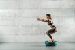 Side view of muscular Caucasian woman in sportswear and with ponytail doing squat endurance on bosu ball. In background wall.