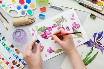 Watercolor drawing - pink cherry flowers - and artistic equipment on desk. Top view. Painter drawing at working place.