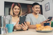 © tirachard - Attractive young Asian couple distracted at table with newspaper and cell phone while eating breakfast. Excited young Asian couple surprised by unbelievably good news, happy family amazed by internet.