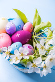 Fototapeta Storczyk - Closeup set of decorated colorful Easter eggs in basket with white spring flowers on light blue background