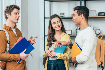 Three happy multicultural students holding notebooks and books and looking at each other