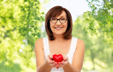 health, charity and valentine's day concept - portrait of smiling senior woman holding red heart over green natural background