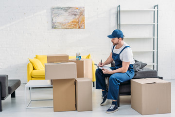 Sticker - handsome mover writing in clipboard while sitting near cardboard boxes in living room