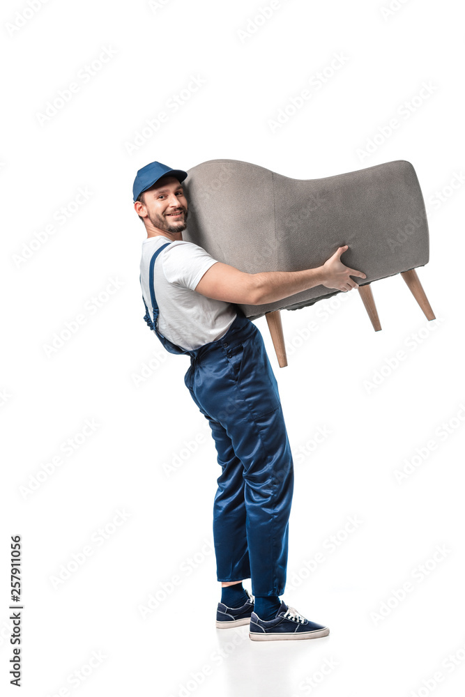 handsome mover in uniform looking at camera while carrying grey armchair isolated on white - obrazy, fototapety, plakaty 