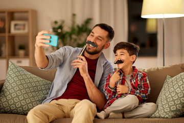 Wall Mural - people, family and technology concept - happy father and little son with mustaches party props taking selfie by smartphone sitting on sofa at home in evening