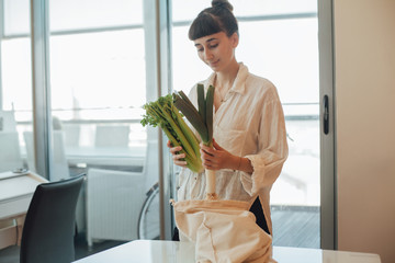 Happy young brunette girl with a tote blank cotton bag for shopping putting fresh green vegetables, leek and greens on the kitchen table. Reusable grocery bag, no plastic, for sustainable shopping.