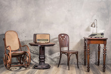 Vintage Loft Room With Antique Rocking Chair, Broadcast Radio, Old Typewriter And Lamp On Oak Wooden Desk Front Concrete Wall Background With Shadows. Retro Style Filtered Photo