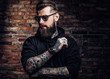 © Fxquadro - A stylish tattooed guy in a black hoodie and sunglasses. Studio photo against a brick wall