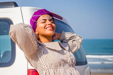 young african american woman in a purple scarf on her head enjoying the summer day next to the car sunny beach and ocean in the background. indian female model in dress resting in car near sea