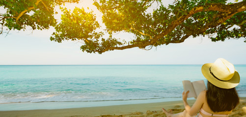 Wall Mural - Women sit and reading a book under the tree at seaside. Back view of sexy Asian woman with straw hat relaxing and enjoying holiday at tropical paradise sand beach. Summer vacation. Summer vibes.