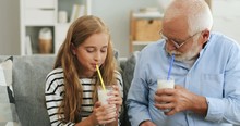 Portrait Of The Cheerful Granfather In Glasses And Cute Granddaughter Drinking Milk Or Milkshakes With Straws On The Couch In The Living Room.