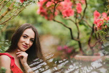 Young beautiful caucasian woman in glass greenhouse among colorful azalea flowers. Art portrait of a long-haired girl wearing a romantic red dress.