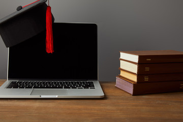 Wall Mural - Laptop with blank screen, books and academic cap with red tassel on wooden surface isolated on grey
