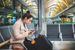 © Daniel Rodriguez - Business woman at the airport sitting waiting for her flight looking at the cell phone. Girl late for distressed plane flight at the international airport. Person sitting with suitcase on vacation sca