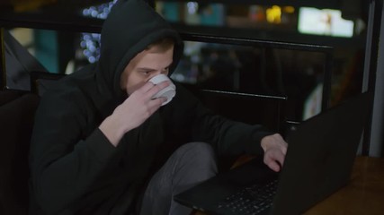 Wall Mural - lockdown of young hooded man sitting in bar at computer, typing on keyboard and drinking hot tea