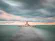 view of a lighthouse in the middle of the sea at sunset along stone walkway submerged in the water and beaten by the waves in long exposure