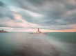 view of a lighthouse in the middle of the sea at sunset along stone walkway submerged in the water and beaten by the waves in long exposure