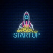 Glowing neon sign of business project startup. Business symbol as a flying rocket starting from coins in neon style