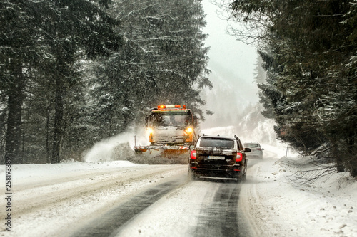 Fotomural Driving on forest road through snowstorm, orange maintenance plough truck coming opposite way, view from car behind