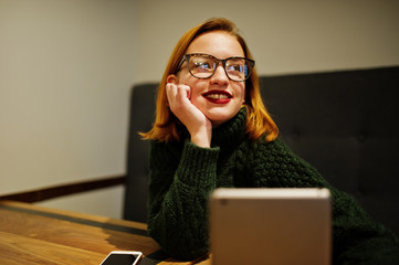 Cheerful young beautiful redhaired woman in glasses, wear on green warm wool sweater,  using her touchpad tablet while sitting at working place on cafe.