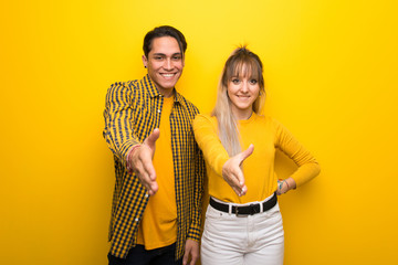 Wall Mural - Young couple over vibrant yellow background shaking hands for closing a good deal