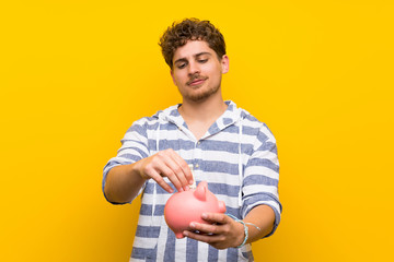 Wall Mural - Blonde man over yellow wall taking a piggy bank and happy because it is full