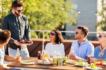 leisure and people concept - happy party host offering meat to his friends at bbq on rooftop in summer